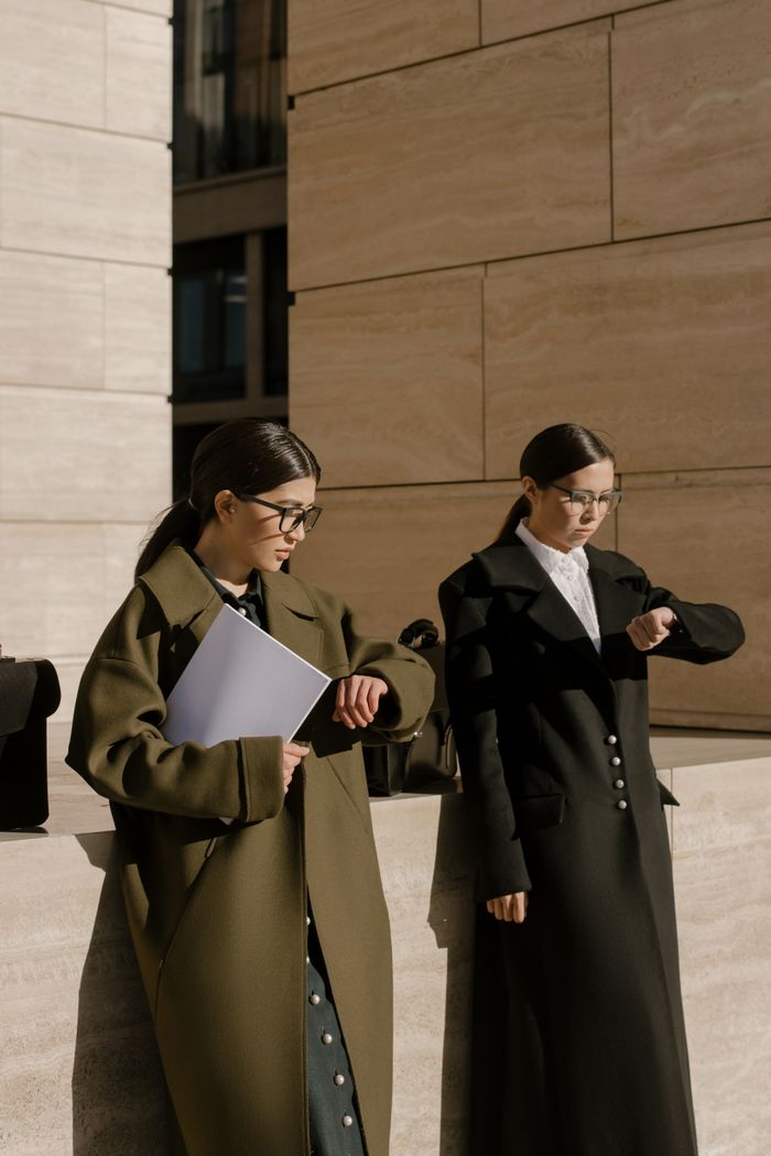 why-choose-us Two fashionable women in long coats checking time under sunlight outdoors.